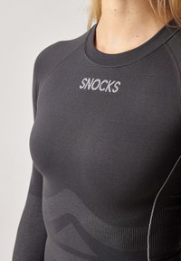 Black long-sleeve athletic top featuring a high neck, textured fabric, and subtle wave pattern. "SNOCKS" logo in silver on the chest.
