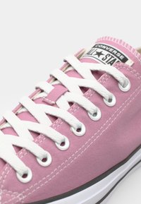 Pink canvas sneaker with white laces, circular metal eyelets, and a black and white logo patch on the tongue. White rubber sole.