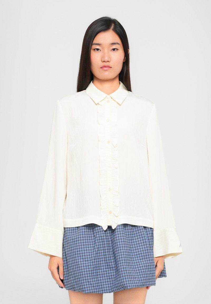 Valge tekstuuriga pluus, millel on volangiline nööpide rivi ja pikad kellukese varrukad, koos sinise ruudulise seelikuga.