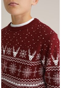 Burgundi színű kötött pulóver fehér geometriai mintákkal, rénszarvas motívumokkal és hópelyhekkel. Bordázott nyakkal és texturált anyaggal rendelkezik.