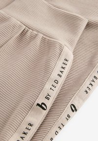 Beige ribbet stof har en tekstureret overflade med et logo bånd, der står "by Ted Baker." Blødt, fleksibelt materiale, egnet til beklædning.