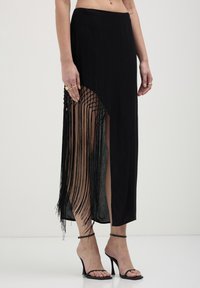Gonna lunga nera con uno spacco laterale e dettagli in frange, caratterizzata da una texture fluida e un design a vita alta. Visibili tacchi neri con strappi sottili.