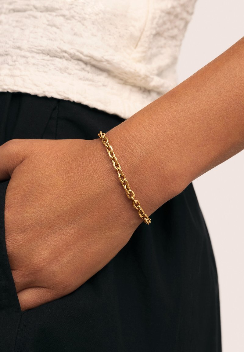 Pulsera de cadena de oro con eslabones entrelazados y un acabado suave y pulido. Lleva puesta en una muñeca sobre un fondo negro.
