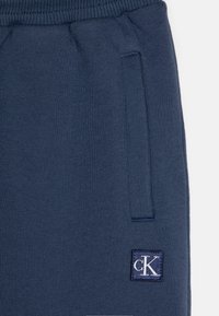 Mørkeblå joggebukser laget av myk fabric, med elastisk midje, sidelommer og et CK-logo-patch i hvitt på en kvadratisk bakgrunn.
