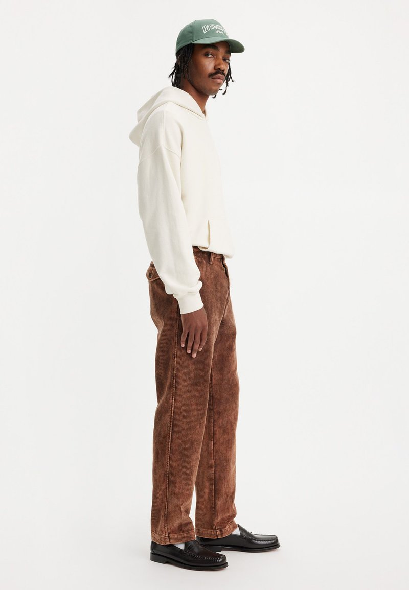 Helle beige Kapuzenjacke, braune Cordhosen und schwarze Slipper. Das Outfit zeichnet sich durch eine lockere Passform und lange Ärmel mit gerippten Bündchen aus; die Mütze ist dunkelgrün.
