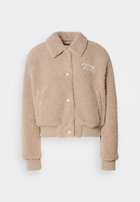 Béžová plyšová skrátená bunda s predným zapínaním na gombíky, rebrovaným lemom a manžetami a vyšitým logom "TOMMY JEANS" na ľavej strane hrude.