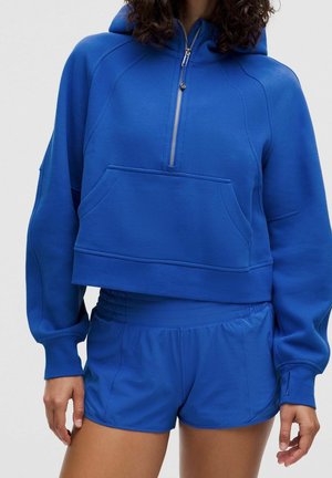Woman wearing a cobalt blue cropped hoodie with a front zipper and matching athletic shorts against a plain background.