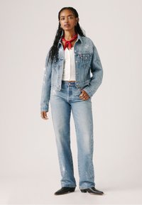 Lichtblauwe denimjas met complexe bloemenborduursels, gecombineerd met bijpassende wijde jeans. Rode bandana om de nek geknoopt en zwarte laarzen.
