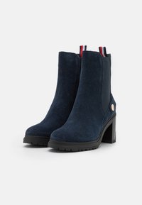 Botines de ante azul marino con un tacón grueso, paneles laterales elásticos y un pequeño acento metálico cerca del tacón. Tiradores rojos y azul marino.