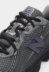 Κοντινό πλάνο ενός γκρι και μοβ αθλητικού παπουτσιού New Balance με μαύρα κορδόνια και σχεδιασμό με διχτυωτό επάνω μέρος.