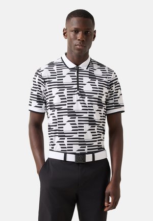 Junger Mann, der ein schwarz-weißes Polohemd mit geometrischem Muster, schwarze Hose und einen weißen Gürtel mit quadratischer Schnalle trägt.