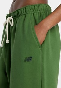Grüne Jogginghose mit elastischem Bund, Kordelzug, Taschen und einem dunkelgrünen New Balance-Logo am linken Oberschenkel.