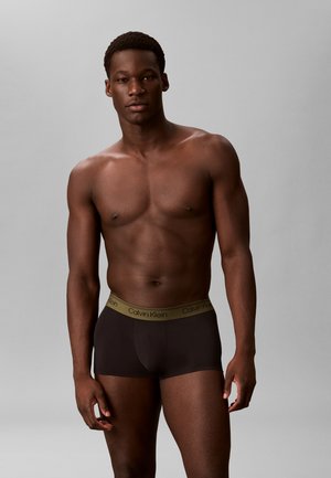 Homme musclé à la peau foncée, torse nu, portant un boxer noir Calvin Klein sur un fond gris uni.