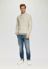 Junger Mann, der nach vorne blickt, trägt einen cremefarbenen Kabelstrickpullover, blaue hochgekrempelte Jeans und weiße Sneaker, steht vor einem einfarbigen Hintergrund.