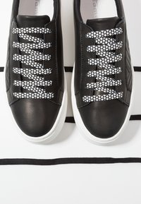 Paire de baskets en cuir noir avec semelles blanches et lacets à motif noir et blanc, posées sur une surface blanche avec des lacets noirs.