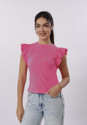 Νεαρή γυναίκα που χαμογελά, φορώντας φούξια αμάνικη μπλούζα με βολάν στους ώμους και ανοιχτόχρωμο τζιν ψηλής μέσης, στέκεται μπροστά σε λευκό φόντο.