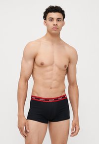 Boxers noirs ajustés avec une ceinture élastique rouge portant l'inscription "HUGO". Le tissu semble lisse, épousant étroitement le corps.