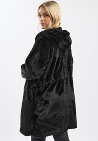 Cappotto in pelliccia sintetica nera con cappuccio, caratterizzato da una vestibilità ampia, cintura elastica e design di lunghezza intermedia. Texture morbida e finitura liscia.