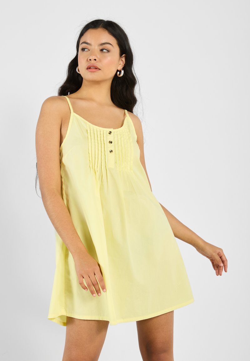 Gelbes ärmelloses Kleid mit lockerer Passform, ausgestattet mit Falten und drei Knöpfen an der Vorderseite. Glattes Material mit minimaler Textur.