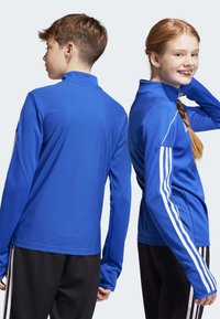 Blaues sportliches Langarmshirt mit strukturiertem Finish, hohem Kragen und weißen Streifen an den Ärmeln, kombiniert mit schwarzen Hosen.