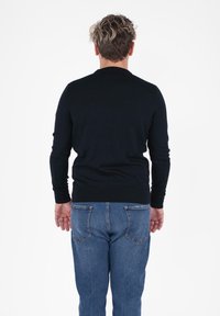 Persona con capelli ricci di spalle, che indossa un maglione nero e jeans blu, in piedi contro uno sfondo bianco uniforme.