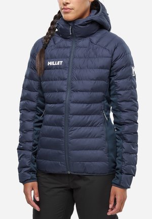 Femme portant une veste matelassée Millet bleu marine avec capuche, fermeture éclair à l'avant et poches latérales, debout avec les mains détendues le long du corps.