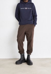 Persona vistiendo una sudadera de color azul marino con el texto "GANT 1949", pantalones jogger marrones y botas de tobillo gruesas negras, de pie contra una pared blanca.