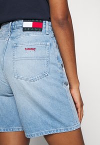 Pantalones cortos de denim azul claro con detalle en el bolsillo trasero, que presentan un bordado en rojo de "tommy jeans" y una cinturilla con marca. Textura suave y diseño casual.