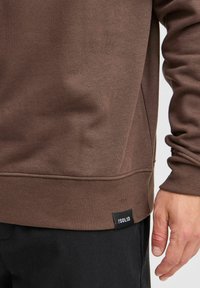 Sweatshirt marron avec des poignets et un ourlet côtelés, comportant une étiquette en couture latérale portant le label "ISOLID". Texture de tissu douce observée à la surface.