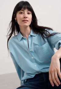 Lichtblauw gestreept shirt met een knoopsluiting, kraag en een borstzak. Het materiaal lijkt ademend en zacht, met lange mouwen.