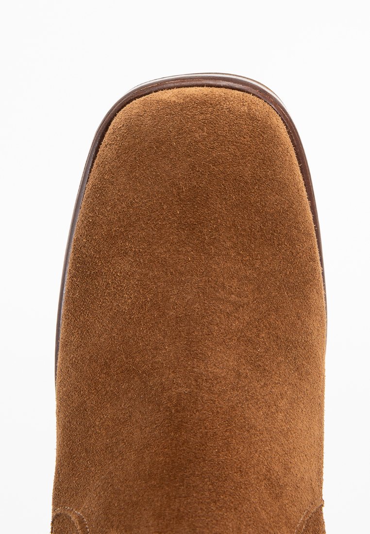 Bottines en daim marron avec une pointe arrondie et des accents en cuir lisse le long des bords. Surface texturée et détails cousus sont visibles.