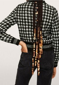 Cardigan en maille à carreaux noir et blanc avec un design ajusté, un matériau texturé et une bordure noire contrastante, associé à un jean noir et des cheveux ornés de perles.