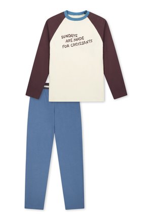 Ragazzo sorridente con pantaloni blu indica una maglietta crema con maniche bordeaux, il testo dice "Sundays are made for croissants," a piedi nudi su sfondo bianco.