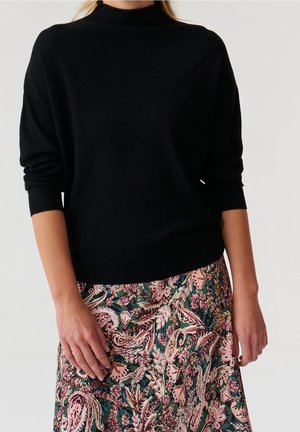 Femme portant un pull noir à col roulé et manches longues ainsi qu'une jupe taille haute à motifs floraux roses, verts et bordeaux.
