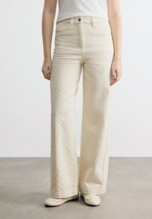 Isik, kes kannab kõrge vöökohaga kreemikaid laiapõlve pükse peene lillemustriga ning valgeid lamedaid jalanõusid, seistes hallil põrandal.