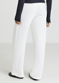 Pantalon blanc à jambes larges avec une texture lisse. Il présente un design épuré, une coupe ajustée, et des chaussures noires contrastantes avec des accents ronds.