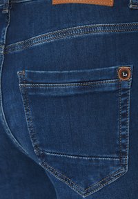 Ciemnoniebieskie dżinsy z pomarańczowymi przeszyciami, tylną kieszenią z ozdobnym guzikiem oraz brązową skórzaną naszywką nad paskiem.