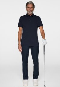 Granatowa koszulka polo z kołnierzykiem, zestawiona z dopasowanymi granatowymi spodniami. Golfista trzyma kij i nosi białą rękawiczkę i buty golfowe.