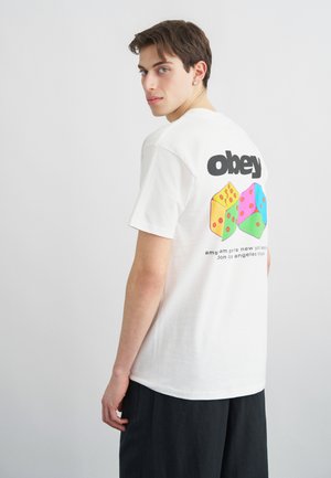 Junger Mann mit kurzen braunen Haaren, trägt ein weißes T-Shirt mit bunten Würfeln und dem Schriftzug "obey" auf der Rückseite, kombiniert mit schwarzen Hosen.