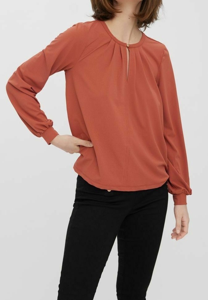 Blouse à manches longues couleur rouille avec un détail en goutte d'eau au niveau du col et des poignets froncés, associée à un pantalon noir ajusté. Tissu au toucher lisse.