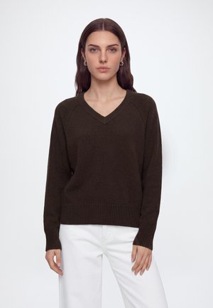 Femme aux longs cheveux bruns portant un pull en V marron foncé et un pantalon blanc, debout devant un fond clair uni.