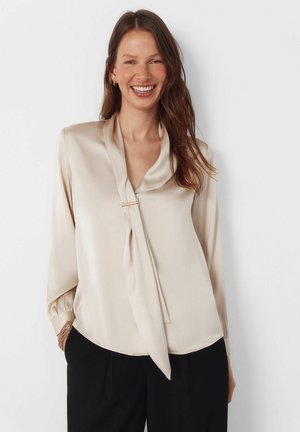 Beige Satinbluse mit Bindeschleife am Ausschnitt, langen Ärmeln und elastischen Manschetten; mit einem goldfarbenen Akzent am Kragen für zusätzliche Details.