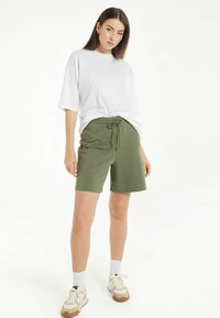 Biała, oversize'owa koszulka z podwiniętymi krótkimi rękawami, zestawiona z oliwkowymi szortami na sznurki. Model nosi białe skarpetki i beżowe sportowe buty.