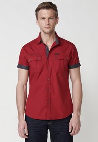 Camisa roja de manga corta con botones y cuello y puños en gris. Presenta dos bolsillos en el pecho y un ajuste relajado. Tejido suave y ligero.