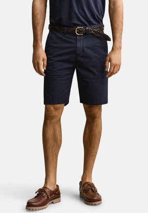Mann trägt marineblaue Shorts, einen dunklen geflochtenen Gürtel und braune Leder-Bootsschuhe, steht vor einem schlichten Hintergrund.