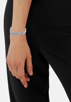 Bracelet en argent avec le mot "croire" en police cursive, posé sur un bracelet texturé blanc. Main sur fond de vêtement noir.