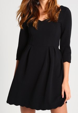 Vrouw met een zwarte, gestructureerde jurk met driekwartmouwen, een V-hals, een aangesloten taille en een geschulpte zoom.