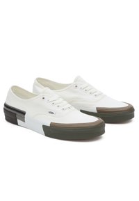 White canvas sneakers with a low-top design, featuring brown and green accents on the rubber sole. Includes six eyelets and textured outsole.