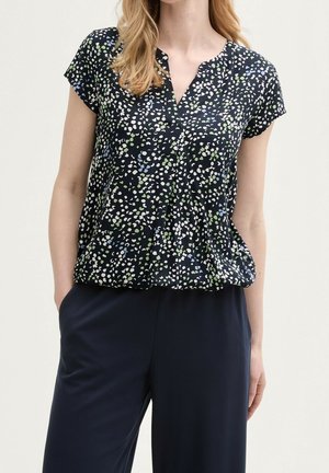 Blouse bleu marine avec un col en V, manches courtes, présentant un motif de taches blanches, vertes et bleues. Tissu doux, coupe ample.