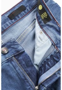 Dżinsy z jasnoniebieskim wnętrzem, czerwonymi przeszyciami, metalowym guzikiem i zamkiem błyskawicznym. Etykiety zawierają napis "Flexcity" oraz ekologiczne przesłanie.